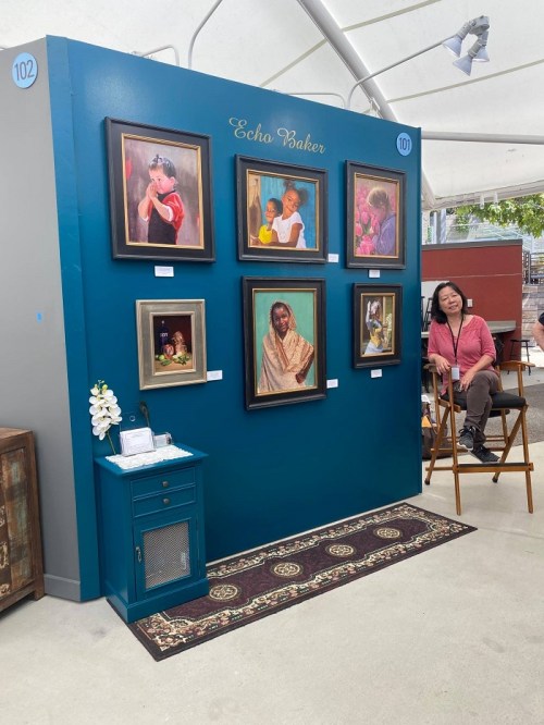 A gallery wall featuring framed portraits by Echo Baker, with an artist seated nearby.