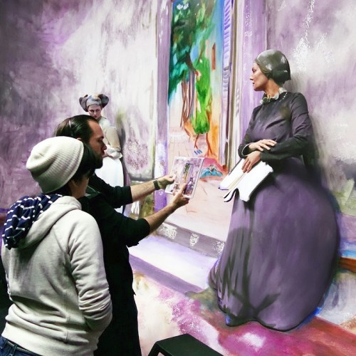 Two people examine a painting of a woman in a gray dress, with another figure in the background, in an art gallery setting.