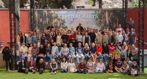A large group of people poses together outdoors at the Festival of Arts, with a colorful mural in the background.