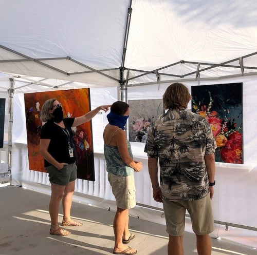 Three people view colorful floral paintings in a tented art display, with one person pointing at the artwork.