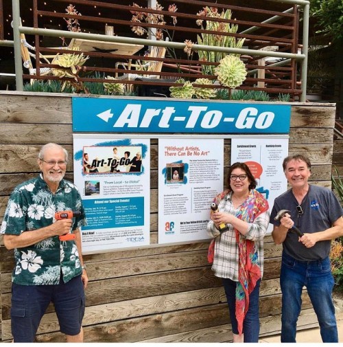 Three people stand in front of an "Art-To-Go" sign, holding tools and smiling, with plants in the background.
