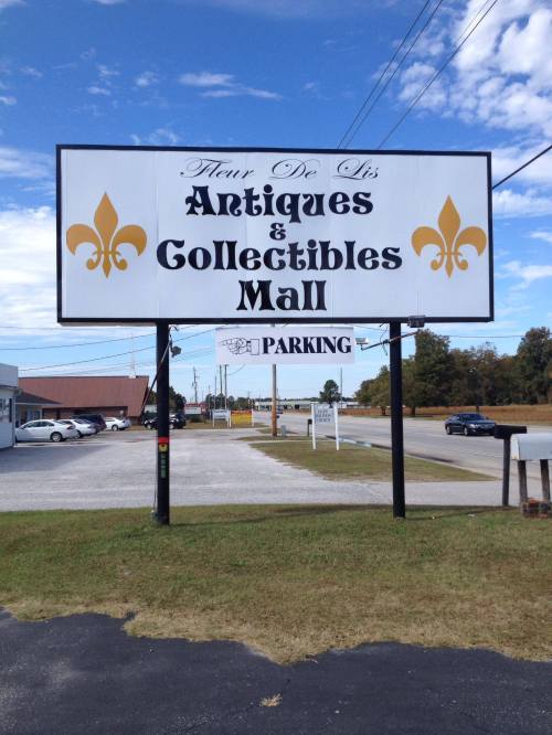 Sign for "Fleur De Lis Antiques & Collectibles Mall" with a parking area, set against a blue sky.