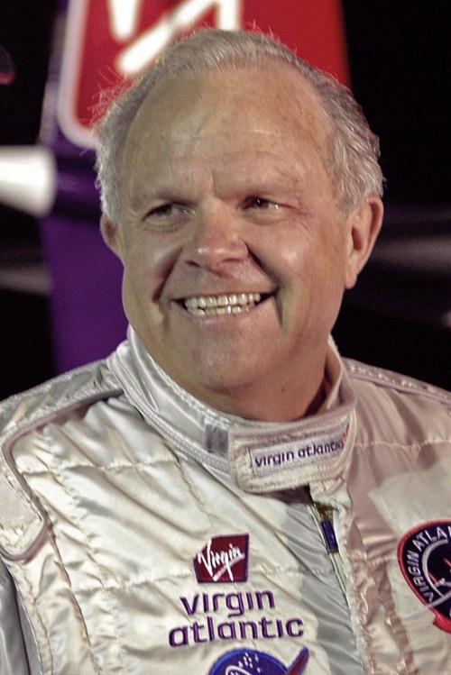 A smiling man in a silver flight suit with Virgin Atlantic logos, standing in front of an aircraft.