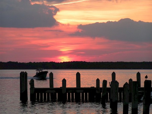 A boat glides past a pier at sunset, with vibrant pink and orange hues reflecting on the water.