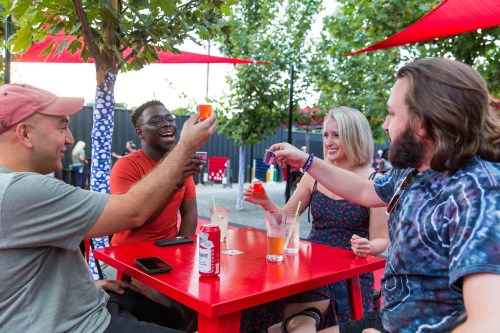 A group of four friends cheers with colorful drinks at a vibrant outdoor setting, surrounded by trees and red canopies.