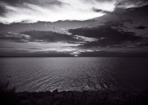 Black and white image of a calm lake at sunset, with dramatic clouds reflecting on the water's surface.