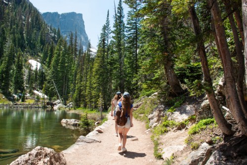Two people walk along a dirt path by a serene lake, surrounded by tall trees and mountains in the background.
