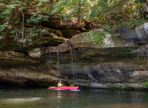 A person in a red kayak paddles near a rocky cliff with a small waterfall and lush greenery above.