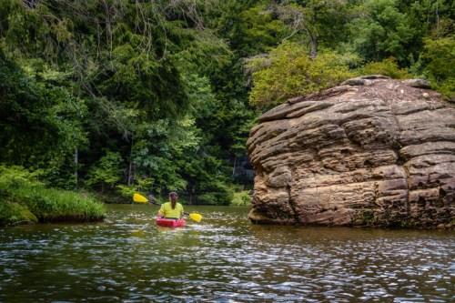 A person kayaking on a calm river surrounded by lush greenery and a large rock formation.