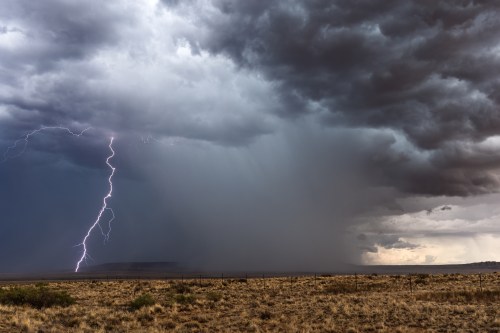 A dramatic storm scene with dark clouds, heavy rain, and a lightning strike over a dry landscape.