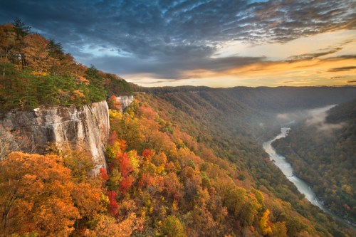A scenic view of a river winding through colorful autumn foliage and cliffs under a dramatic sky at sunset.