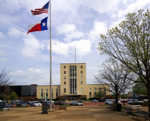 A government building with American and Texas flags, surrounded by trees and parked cars under a cloudy sky.