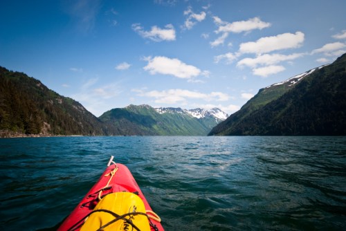 A kayak on a serene lake, surrounded by lush green mountains and snow-capped peaks under a clear blue sky.