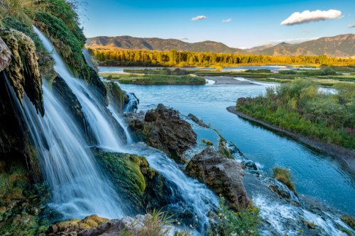 A scenic view of a waterfall cascading into a river, surrounded by lush greenery and mountains in the background.