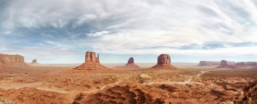 A panoramic view of Monument Valley, featuring iconic red rock formations under a cloudy sky.