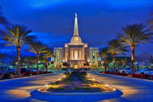 A beautifully lit temple at dusk, surrounded by palm trees and landscaped gardens, with a tall spire reaching into the sky.