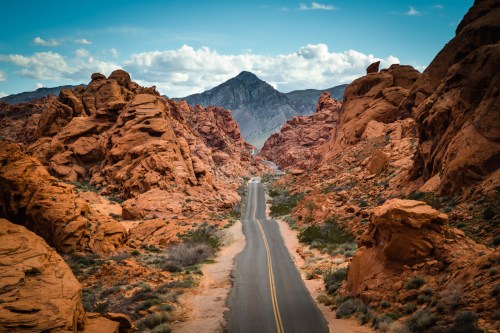 A winding road cuts through vibrant red rock formations under a blue sky with scattered clouds.