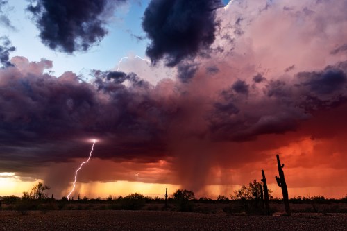 Dramatic sunset with dark clouds, lightning, and rain over a desert landscape with silhouetted cacti.