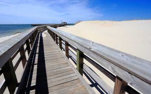 A wooden boardwalk leads to a sandy beach and pier under a clear blue sky.