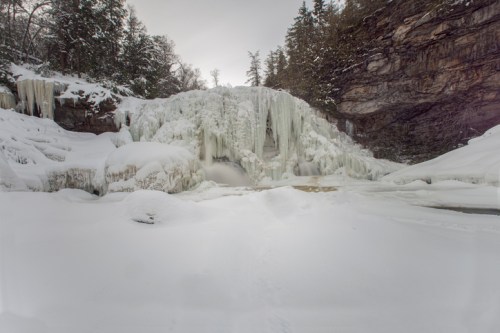 A frozen waterfall surrounded by snow-covered rocks and trees, creating a serene winter landscape.