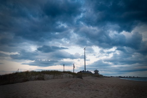 A beach scene with sand dunes, a wind turbine, and a cloudy sky at dusk.