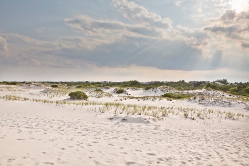 A serene beach landscape with soft white sand, gentle dunes, and a cloudy sky illuminated by rays of sunlight.