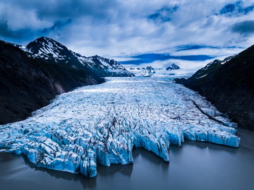Aerial view of a vast glacier flowing into a calm lake, surrounded by rugged mountains and a cloudy sky.
