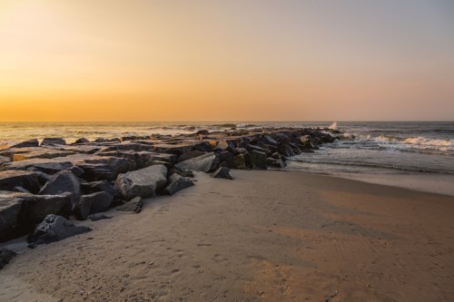 A rocky jetty extends into the ocean at sunset, with gentle waves lapping against the shore.
