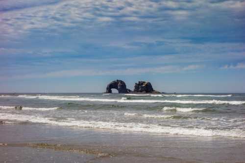 A scenic beach view featuring two rock formations in the ocean under a blue sky with scattered clouds.