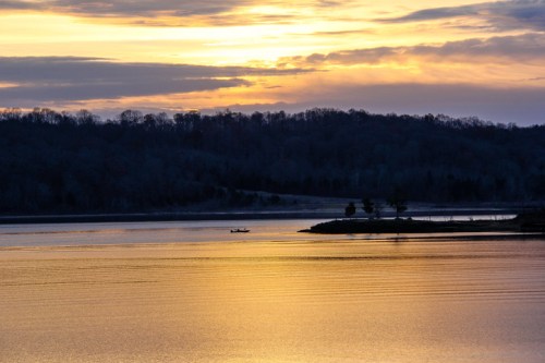 A serene sunset over a calm lake, with a small boat silhouetted against the colorful sky and distant trees.