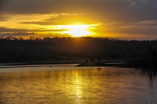 Sunset over a calm lake, with golden reflections on the water and silhouettes of trees in the background.