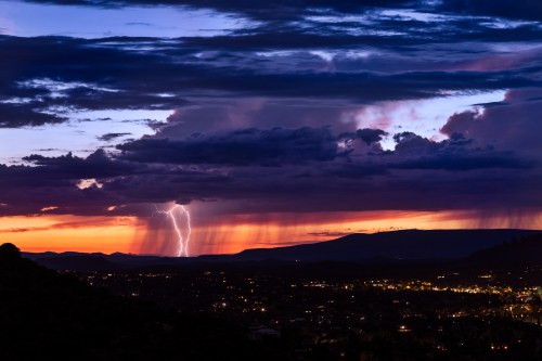 A dramatic sunset with dark clouds, lightning striking in the distance, and city lights twinkling below.