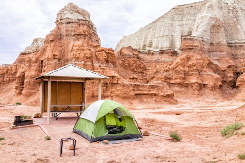 A green tent set up near a picnic shelter in a desert landscape with red rock formations and cloudy skies.
