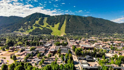 Aerial view of a town surrounded by mountains, with ski slopes visible on the hillside and greenery throughout.