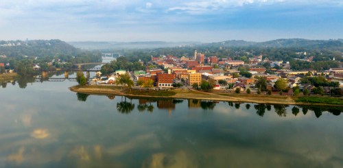 Aerial view of a small town by a river, featuring buildings, trees, and a misty landscape in the background.
