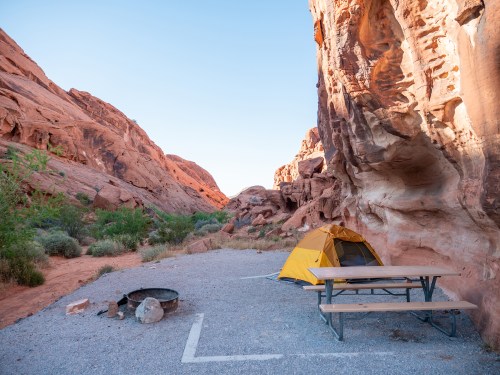 A yellow tent beside a picnic table and fire pit, surrounded by rocky cliffs and desert vegetation in a serene campsite.