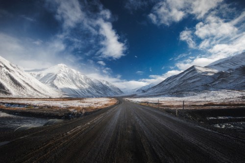 A winding road stretches through snow-capped mountains under a dramatic blue sky with scattered clouds.
