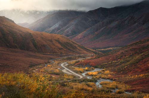 A winding road through colorful autumn mountains, with a river flowing alongside in a serene landscape.