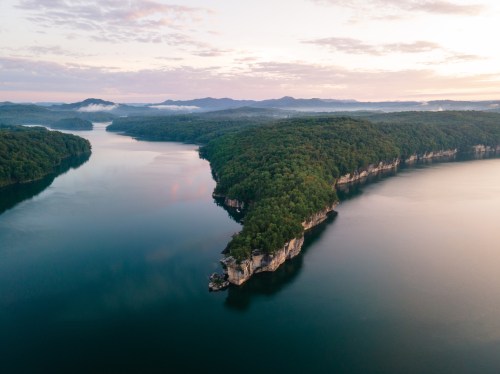 Aerial view of a serene lake surrounded by lush green hills and cliffs under a soft, cloudy sky at sunrise.
