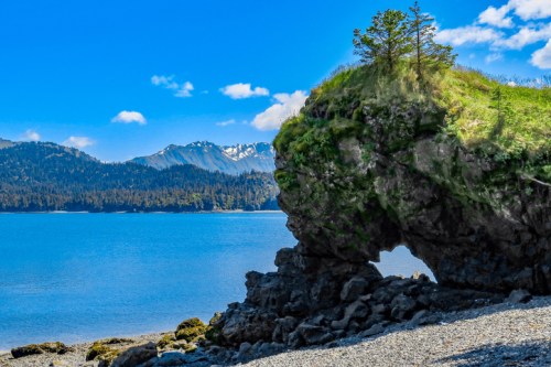 A rocky shoreline with a grassy hill, overlooking a calm blue sea and distant snow-capped mountains under a clear sky.