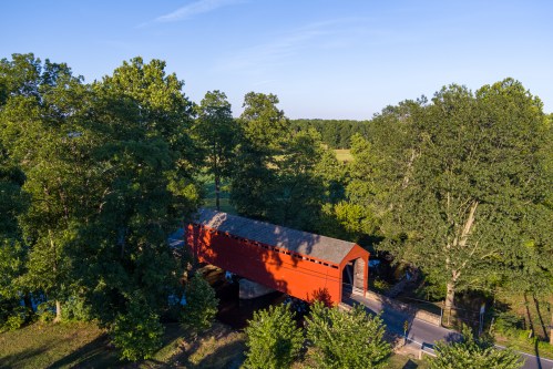 Aerial view of a red covered bridge surrounded by lush green trees and a clear blue sky.