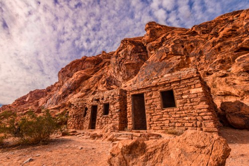 A weathered stone structure nestled against a rocky cliff under a cloudy sky. Desert vegetation surrounds the area.