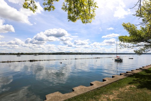 A serene lake scene with a sailboat, calm water, and fluffy clouds under a blue sky, framed by green trees.