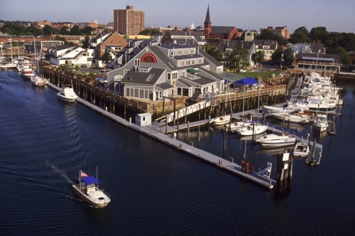 Aerial view of a marina with boats, a waterfront building, and a city skyline in the background.
