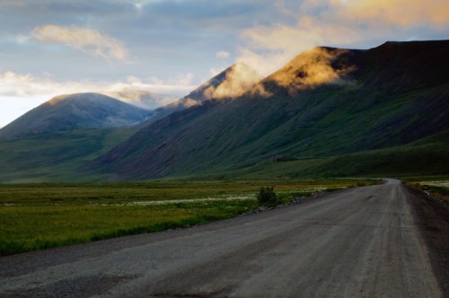 A winding dirt road leads through lush green hills and mountains under a cloudy sky at sunset.