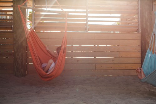 A person relaxes in a hammock under sunlight, with wooden slats in the background and sand beneath.