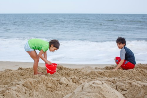 Two children play on a beach, one digging in the sand with a red bucket while the other sits nearby.