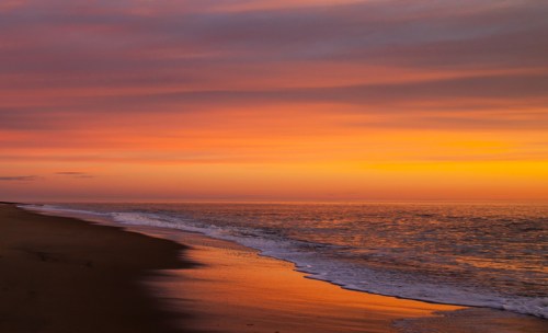 A serene beach at sunset, with vibrant orange and purple skies reflecting on calm waters and wet sand.