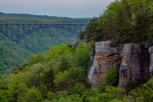 A scenic view of a lush green valley with a bridge spanning across it, framed by rocky cliffs and trees.