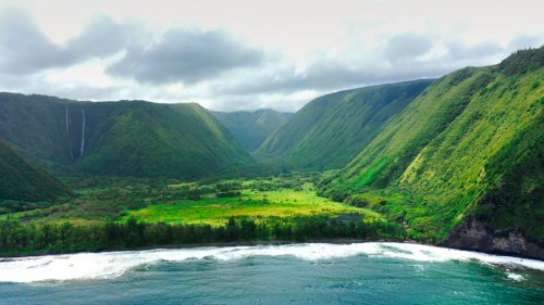 Lush green valleys and mountains meet the ocean under a cloudy sky, with a waterfall cascading in the distance.